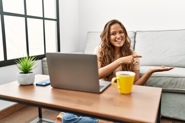 Beautiful hispanic woman using computer laptop at home amazed and smiling to the camera while presenting with hand and pointing with finger.
