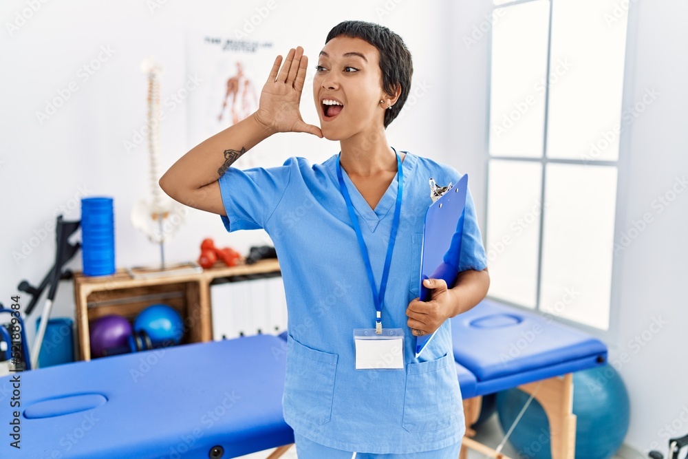 Wall mural Young hispanic woman with short hair working at pain recovery clinic shouting and screaming loud to side with hand on mouth. communication concept.