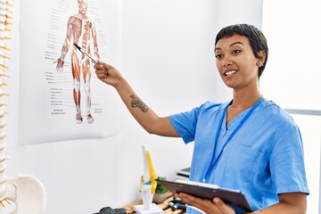 Young hispanic woman wearing physiotherapist uniform pointing to vertebral column holding checklist...