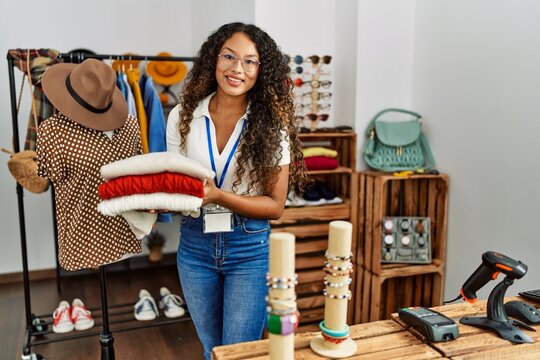 Young latin shopkeeper woman smiling happy holding stack of sweater at clothing store.