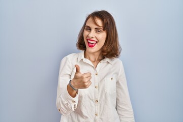Young beautiful woman standing casual over blue background doing happy thumbs up gesture with hand. approving expression looking at the camera showing success.