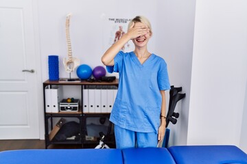 Young physiotherapist woman working at pain recovery clinic smiling and laughing with hand on face covering eyes for surprise. blind concept.