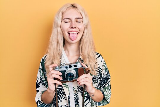 Beautiful caucasian woman with blond hair holding vintage camera sticking tongue out happy with funny expression.