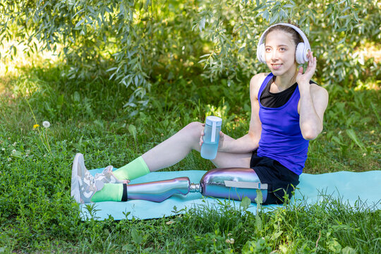 Young beautiful athletic woman with bionic prosthetic leg doing sport exercises in the park outdoors in summer. Concept of active lifestyle of disabled people. Listening to music, drinking water