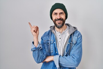 Young hispanic man with tattoos wearing wool cap with a big smile on face, pointing with hand and finger to the side looking at the camera.