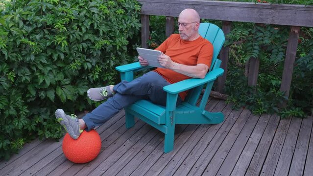 senior man in late 60s is relaxing in an Adirondack chair with a digital tablet on a wooden backyard deck