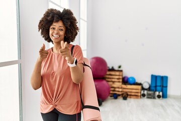 Fototapeta na wymiar African american woman with afro hair holding yoga mat at pilates room pointing fingers to camera with happy and funny face. good energy and vibes.
