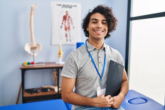 Young hispanic man physiotherapist holding clipboard at rehab clinic