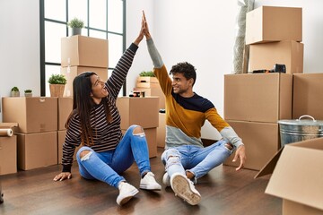 Young latin couple high five with hands raised up sitting on the floor at new home.