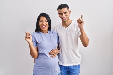 Young hispanic couple expecting a baby standing over background with a big smile on face, pointing with hand finger to the side looking at the camera.