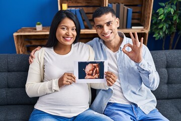 Young hispanic couple expecting a baby sitting on the sofa showing baby ultrasound doing ok sign...