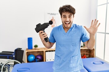 Hispanic physiotherapy man working at pain recovery clinic with muscle gun celebrating victory with happy smile and winner expression with raised hands