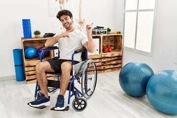 Hispanic man sitting on wheelchair at physiotherapy clinic smiling and looking at the camera pointing with two hands and fingers to the side.