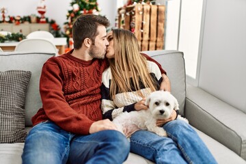 Young hispanic couple sitting on the sofa with dog kissing at home.