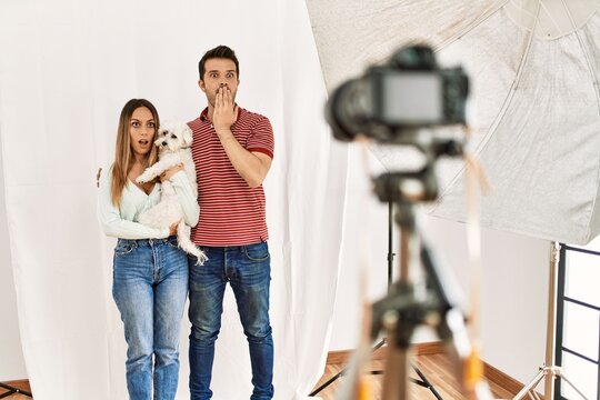 Couple of boyfriend and girlfriend with dog posing as model at photography studio covering mouth with hand, shocked and afraid for mistake. surprised expression