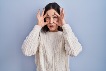 Young brunette woman standing over blue background trying to open eyes with fingers, sleepy and tired for morning fatigue