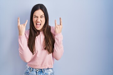 Fototapeta na wymiar Young brunette woman standing over blue background shouting with crazy expression doing rock symbol with hands up. music star. heavy concept.