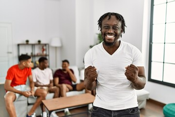 Young african man with friends at the living room very happy and excited doing winner gesture with arms raised, smiling and screaming for success. celebration concept.