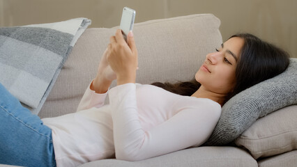 Arabic latino restful lazy young woman lebanese girl freelancer lying on sofa at home relaxing reading sms message on smartphone cell electronic device shopping online make internet order free day off - obrazy, fototapety, plakaty