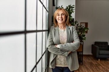 Middle age businesswoman standing with arms crossed gesture leaning on the window at the office.