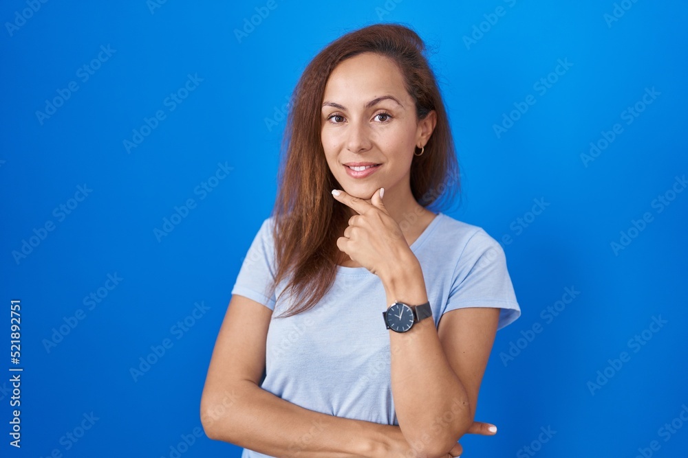 Poster brunette woman standing over blue background looking confident at the camera smiling with crossed ar