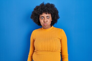 Fototapeta na wymiar Black woman with curly hair standing over blue background puffing cheeks with funny face. mouth inflated with air, crazy expression.