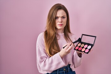 Young caucasian woman holding makeup brush and blush palette skeptic and nervous, frowning upset...