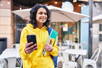 Young latin woman student holding books using smartphone at street