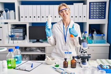 Fototapeta premium Young caucasian woman working at scientist laboratory holding body lotion smiling happy and positive, thumb up doing excellent and approval sign