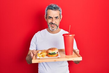 Handsome middle age man with grey hair eating a tasty classic burger with fries and soda clueless and confused expression. doubt concept.