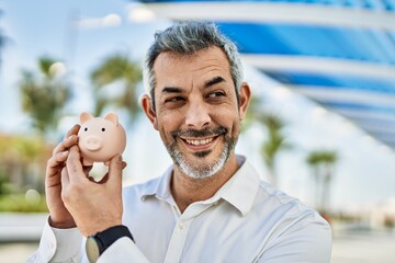 Middle age grey-haired man smiling happy holding piggy bank at the city.