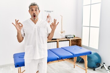Middle age hispanic therapist man working at pain recovery clinic looking surprised and shocked doing ok approval symbol with fingers. crazy expression