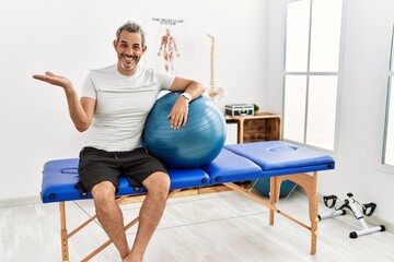 Middle age hispanic man at pain recovery clinic holding pilates ball pointing aside with hands open palms showing copy space, presenting advertisement smiling excited happy