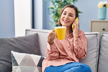 Middle age woman drinking coffee and talking on the smartphone at home