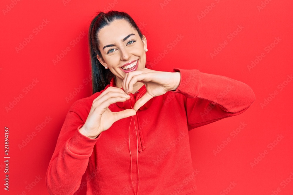 Poster beautiful woman with blue eyes wearing gym clothes and headphones smiling in love showing heart symb