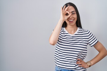 Young brunette woman wearing striped t shirt doing ok gesture with hand smiling, eye looking through fingers with happy face.