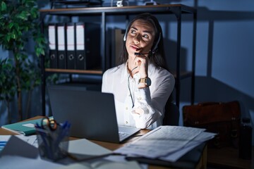 Young brunette woman wearing call center agent headset working late at night with hand on chin...