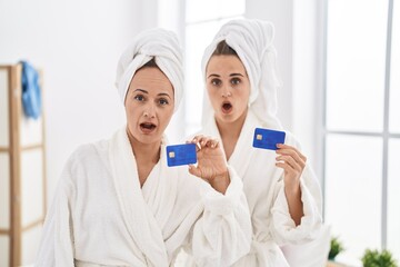 Middle age woman and daughter wearing bathrobe holding credit card scared and amazed with open mouth for surprise, disbelief face