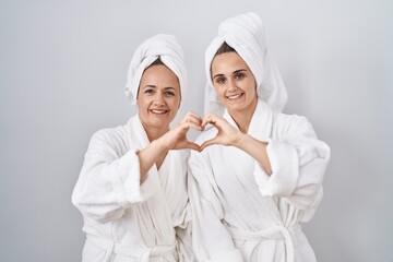 Middle age woman and daughter wearing white bathrobe and towel smiling in love doing heart symbol...
