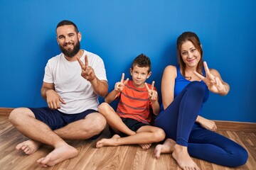 Family of three sitting on the floor at home smiling looking to the camera showing fingers doing...