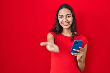 Young brazilian woman using smartphone over red background smiling cheerful offering palm hand giving assistance and acceptance.