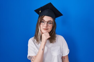 Blonde caucasian woman wearing graduation cap looking confident at the camera with smile with crossed arms and hand raised on chin. thinking positive.