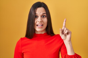 Young hispanic woman standing over yellow background pointing finger up with successful idea. exited and happy. number one.