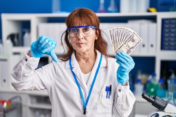 Middle age hispanic woman working at scientist laboratory holding dollars with angry face, negative sign showing dislike with thumbs down, rejection concept