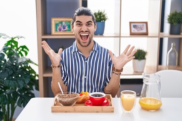 Fototapeta na wymiar Hispanic man with long hair sitting on the table having breakfast celebrating victory with happy smile and winner expression with raised hands