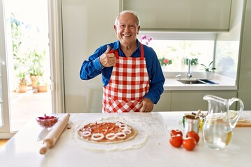 Senior man with grey hair cooking pizza at home kitchen smiling happy and positive, thumb up doing excellent and approval sign
