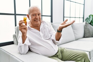 Senior man holding pills smiling cheerful presenting and pointing with palm of hand looking at the camera.