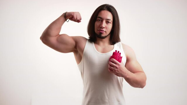 Caucasian muscular man with long dark hair and beard pumping fake anatomical heart in one hand and flexing other arm. Isolated. High quality 4k footage