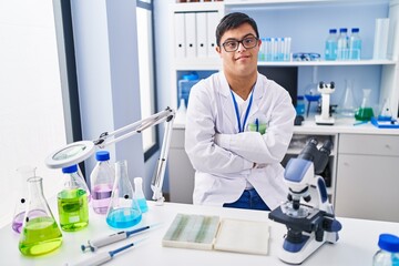 Down syndrome man wearing scientist uniform sitting with arms crossed gesture at laboratory