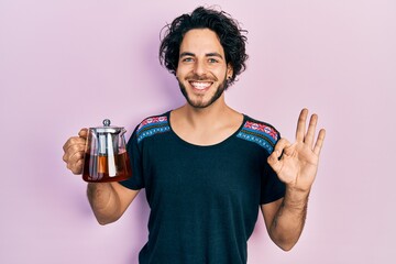 Handsome hispanic man holding traditional tea pot doing ok sign with fingers, smiling friendly gesturing excellent symbol